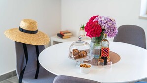 A round dining table with a white tablecloth, a wicker hat, a glass dome with pastries, a vase with flowers, and two bottles. - Apartments Amalija (Konavle)