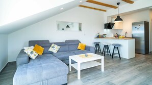 A modern loft apartment with a gray sectional sofa, a white coffee table, and a kitchen area with a stainless steel refrigerator and bar stools. - Apartments Amalija (Konavle)