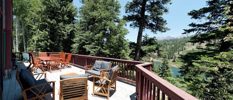 A wooden deck with outdoor furniture, surrounded by tall trees and a view of a lake.