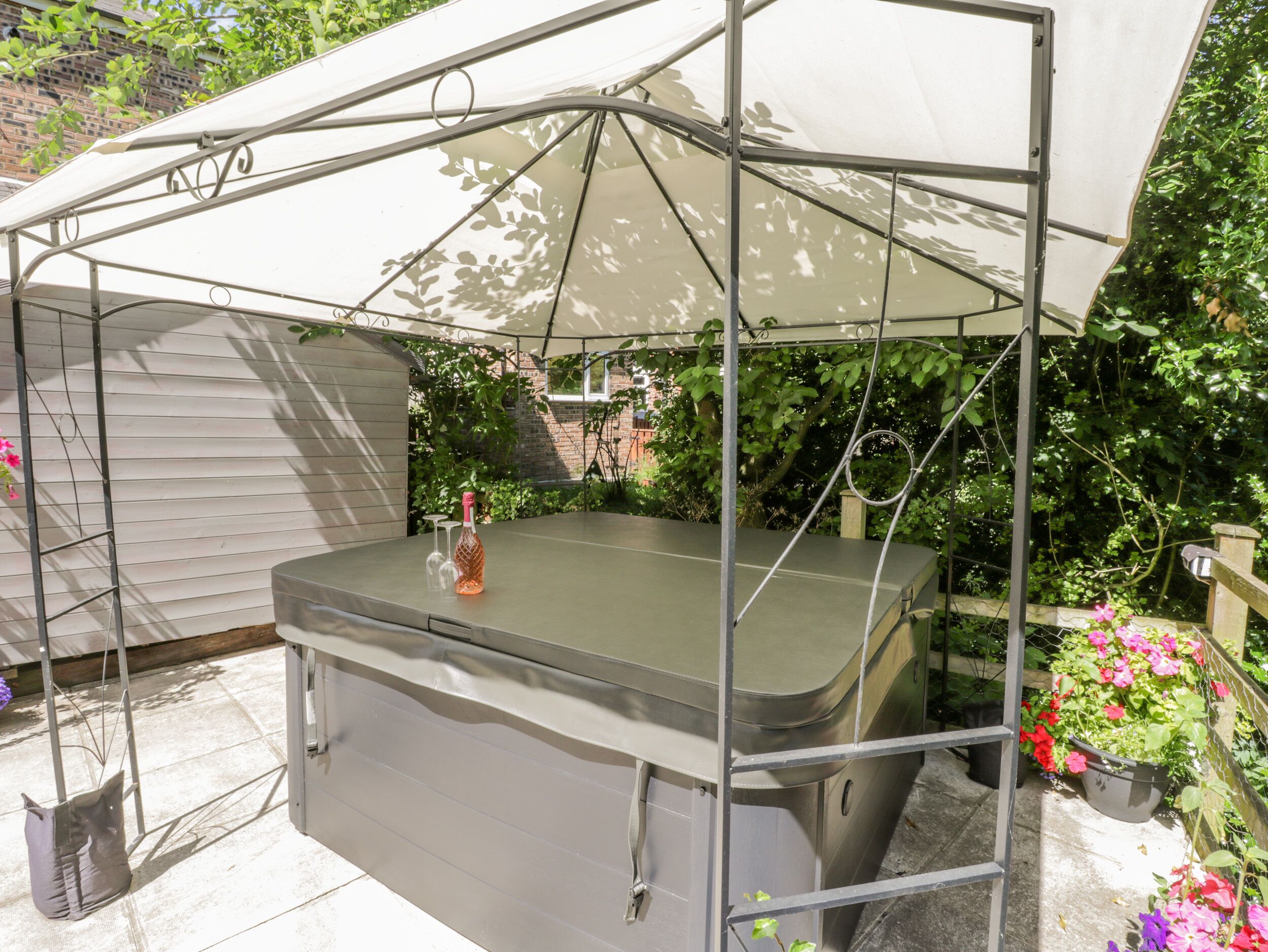 A covered outdoor hot tub with a metal frame and a canopy, surrounded by greenery and a brick wall.