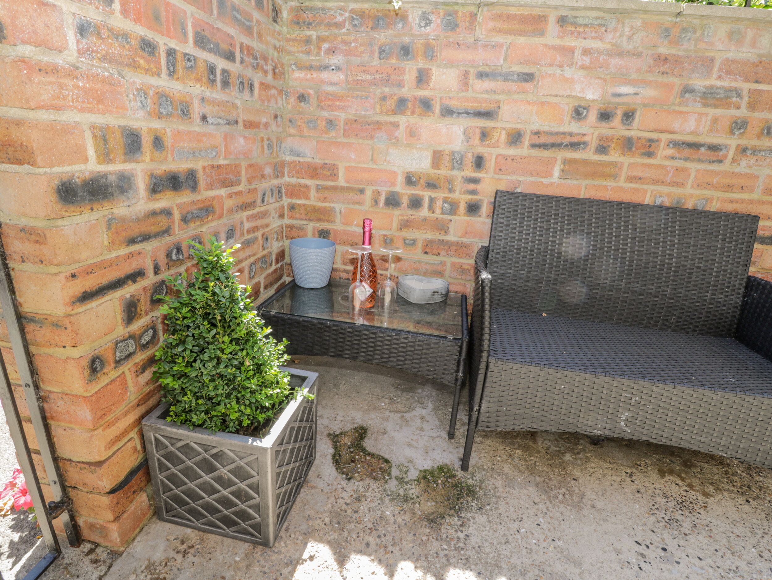 A brick wall, a wicker chair, a small table with a bottle and a bowl, and a potted plant.