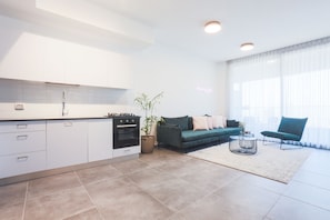 A modern kitchen with white cabinets and a built-in oven, a green sofa with cushions, a round coffee table, and a large window with sheer curtains. - AirTLV - Menachem Begin Apartment (Tel Aviv)