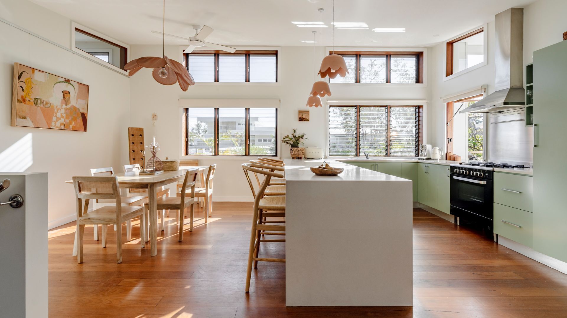 A spacious kitchen with a central island, wooden dining table, and chairs. There are large windows providing natural light and views of the outdoors.