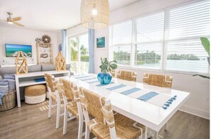 A bright living room with a white sofa, a dining table with wicker chairs, a ceiling fan, and a large window with blinds.