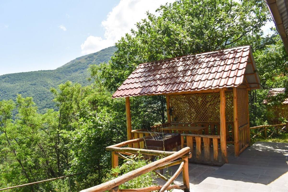 A wooden gazebo with a tiled roof, surrounded by lush greenery and a mountainous backdrop.
