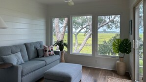 Una sala de estar moderna con un sofá gris, un ventilador de techo y ventanas amplias que ofrecen vistas a un paisaje verde.