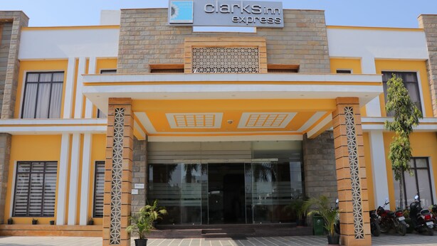 The exterior of a Clark's Inn Express hotel with a modern design, featuring a prominent entrance, a covered area with seating, and a parking area with motorcycles. - Hotel Clarks Inn Express Bharatpur (Bharatpur)