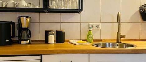 Une cuisine avec un plan de travail en bois, un évier en acier inoxydable et une machine à café. On trouve des placards sous le plan de travail et un réfrigérateur sur la gauche.