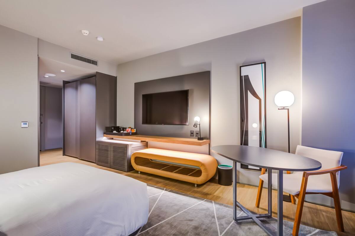 Une chambre d’hôtel moderne équipée d’un lit, d’un coin salon avec une table et des chaises, d’un téléviseur fixé au mur et d’un grand miroir.
