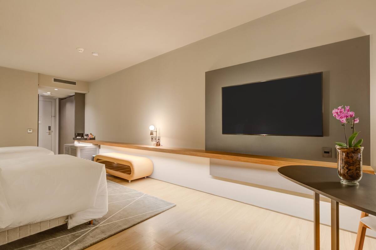 Une chambre d’hôtel moderne avec un grand lit, une table de chevet, un téléviseur à écran plat fixé au mur et une petite plante en pot.