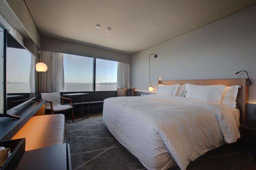 A hotel room with a large bed, a desk, a chair, a TV, and a view of the city through large windows.