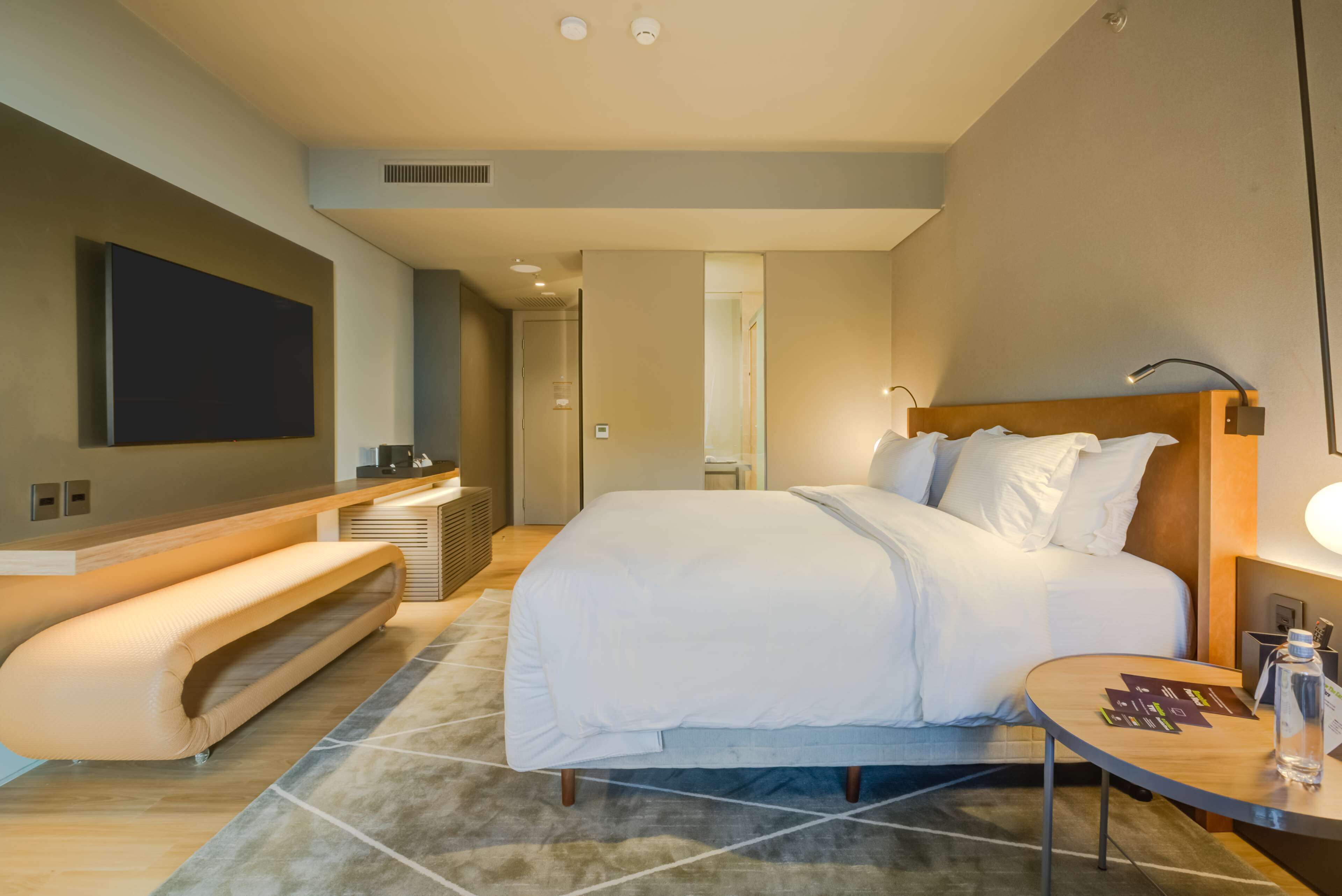 A modern hotel room with a large bed, a bench, a flat-screen TV, a bedside table, and a lamp.