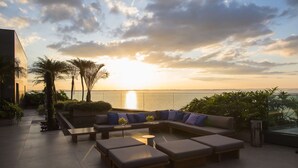 An outdoor seating area with a sofa and benches, overlooking a body of water at sunset.