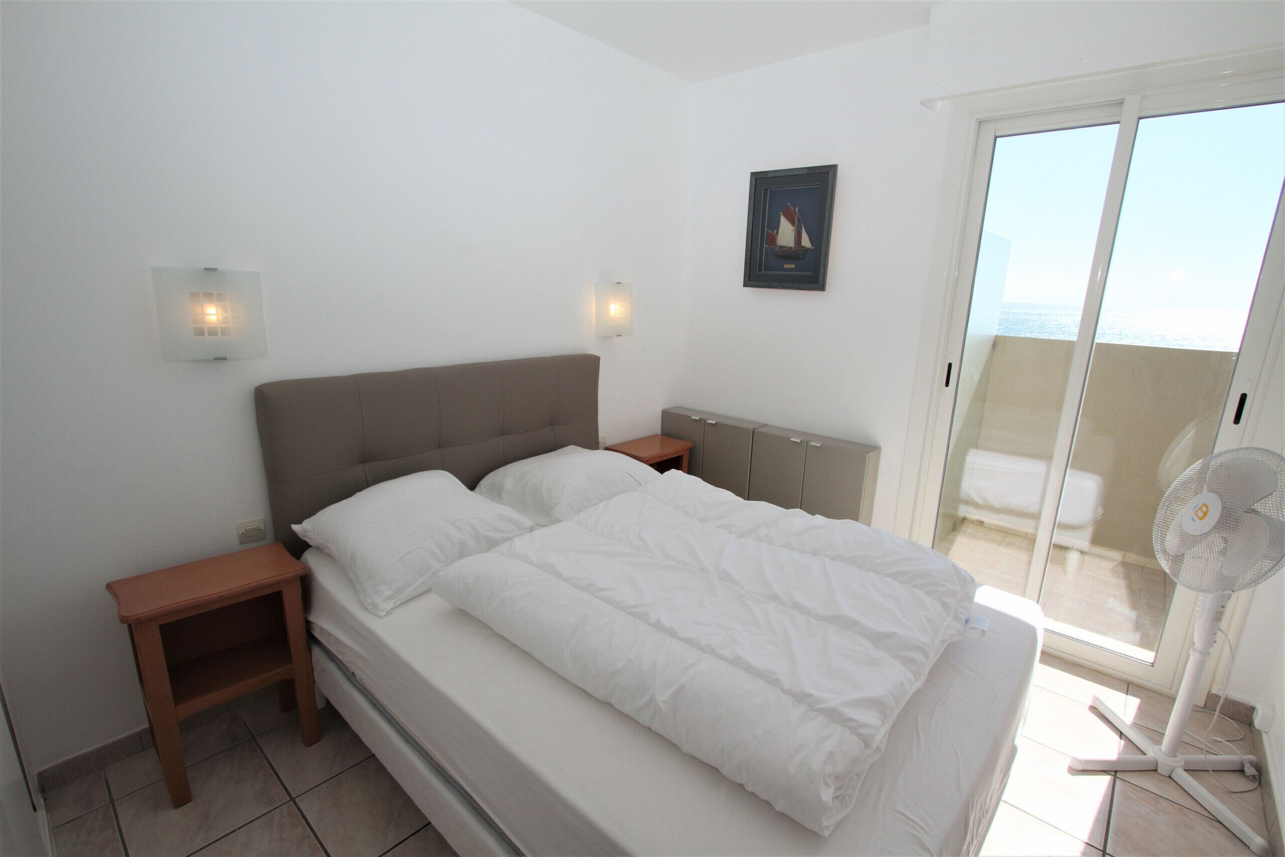 Een slaapkamer met een bed, nachtkastjes, een wandlamp, een lijst met een afbeelding en een balkon met uitzicht.