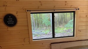 A wooden cabin room with a bed, a window with a view of a forest, and a wall-mounted sign. - Ricketts Glen Fern View Cabin (Benton)