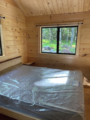 A wooden cabin room with a bed, a window, and a view of a forest. - Ricketts Glen Fern View Cabin (Benton)