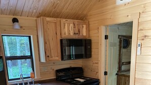 A compact kitchen with wooden cabinets, a black stove, and a microwave. There's a sink with a faucet, a window, and a door leading to another room. A brown leather couch is in the living area. - Ricketts Glen Fern View Cabin (Benton)