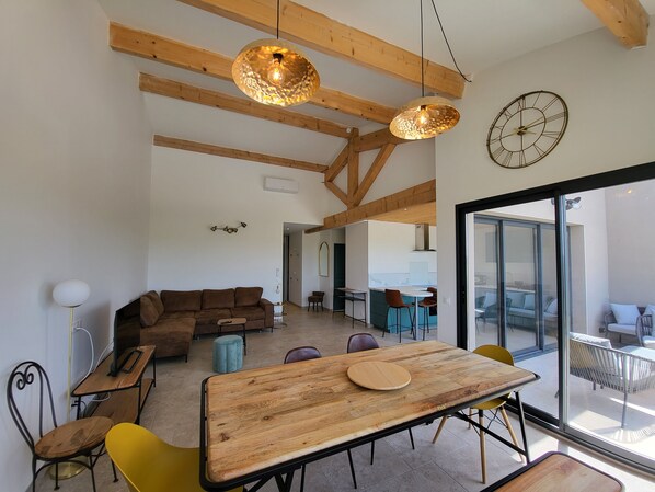 A spacious living area with a wooden ceiling, a dining table with chairs, a sofa, and a large window offering a view of the outdoors.