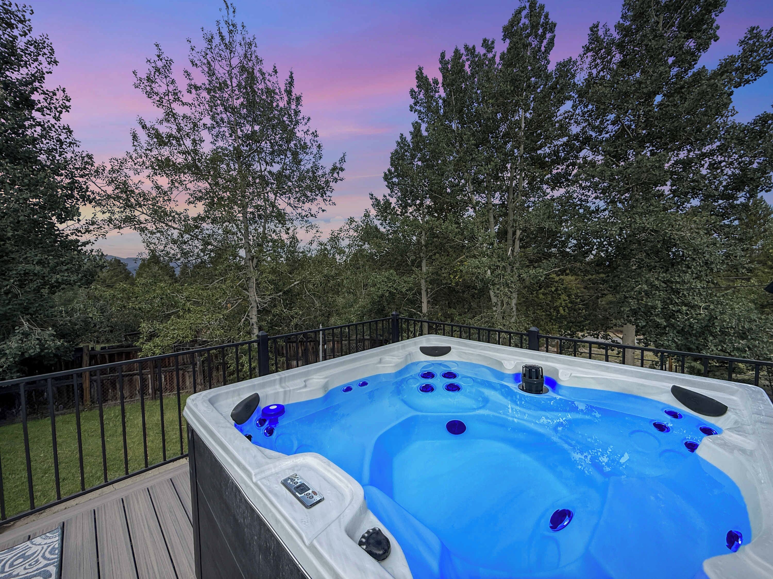 A hot tub on an outdoor deck with a wooden railing, surrounded by trees and a clear sky.