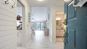 Un pasillo que conduce a una cocina y zona de comedor, con paredes blancas, una puerta azul y un ventilador de techo.
