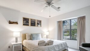 Un dormitorio con cama, dos lámparas de noche, ventilador de techo, ventana con cortinas y tres cuadros en la pared.