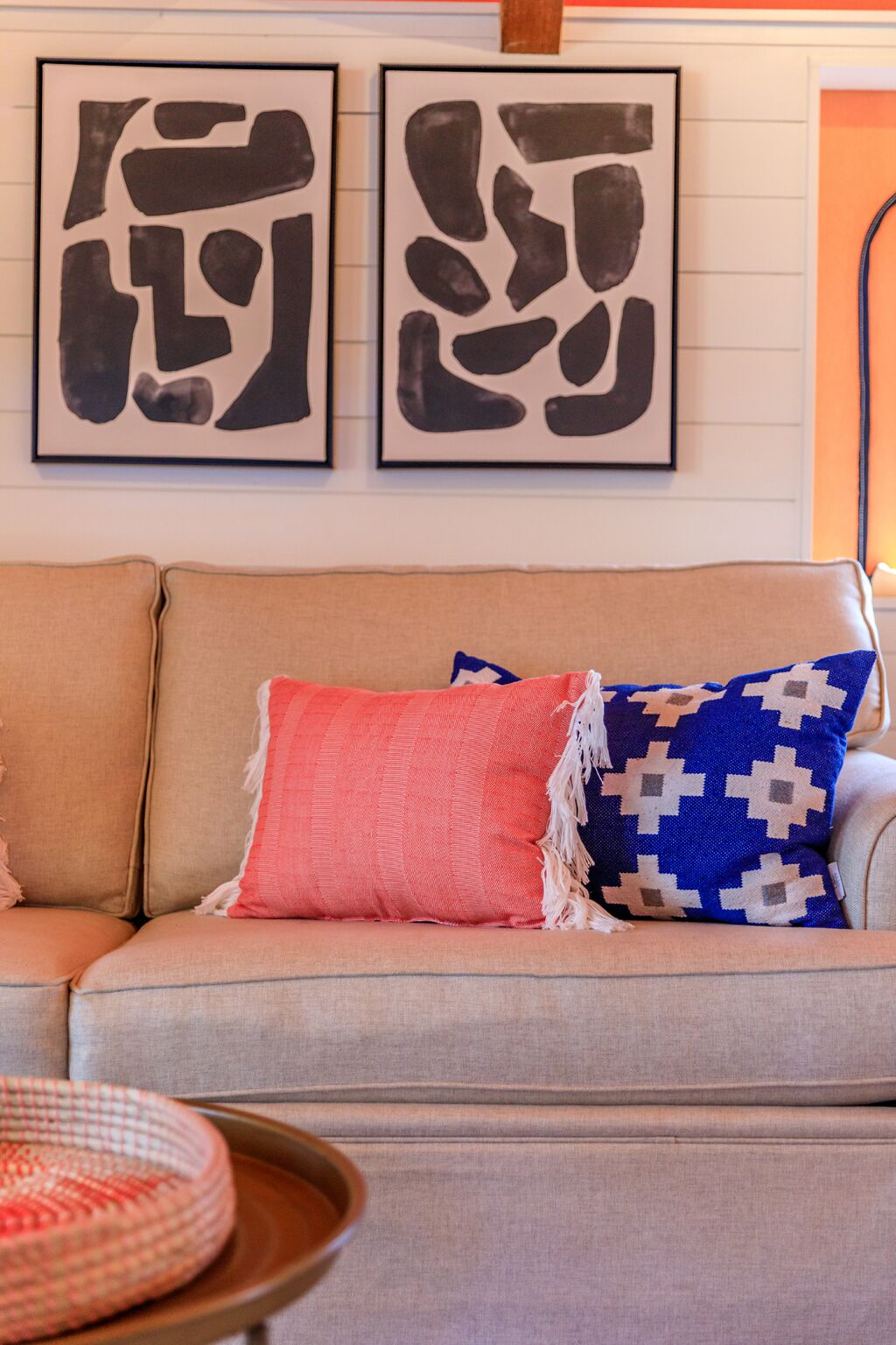 Un sofá beige con dos almohadones decorativos, uno rojo y otro azul con estampado geométrico, y dos obras de arte abstracto en blanco y negro en la pared de arriba.