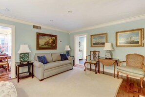 A living room with a sofa, two chairs, a small table, and a lamp. There are paintings on the wall and a door leading to another room. - Liberty Garden, Historic | Cozy | Free Parking (Savannah)