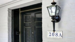 Eine schwarze Tür mit einer weißen Nummernschild, auf der „208A“ steht, an einer weißen Ziegelwand angebracht, begleitet von einer schwarzen Wandleuchte.