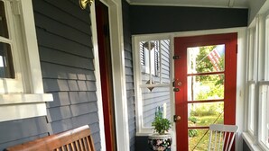 A wooden bench and two white chairs on a porch with a red door and windows.