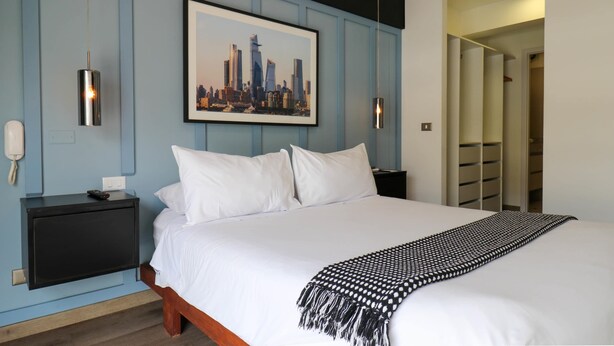 A hotel room with a bed, a framed cityscape picture, a black nightstand, a white bedside table, and a closet with sliding doors.