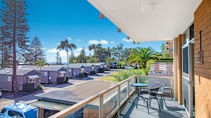 A balcony with a view of a row of cabins, a palm tree, and a clear blue sky. - Crystal Waters 2, 2 Munster Street (PORT MACQUARIE)