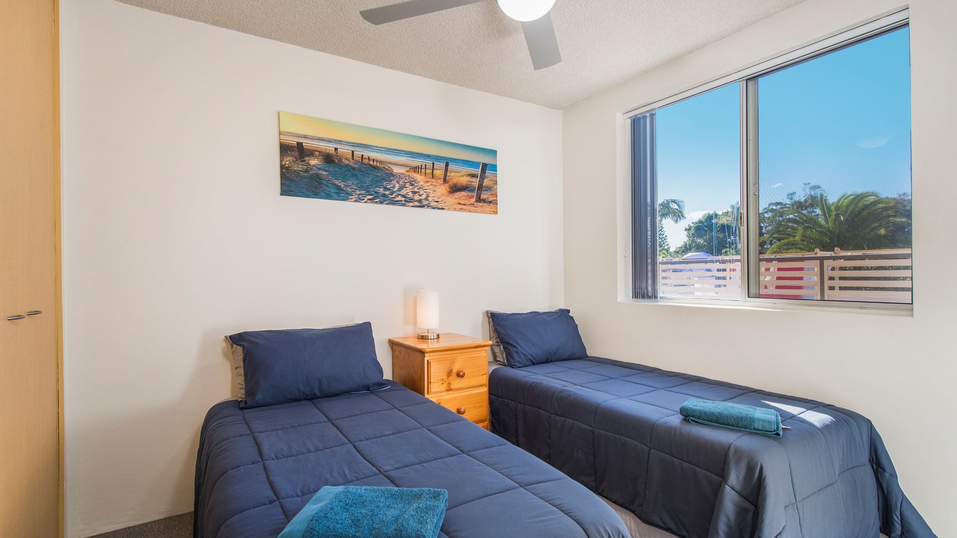 A bedroom with two beds, a nightstand, a ceiling fan, a window with a view, and a painting on the wall.