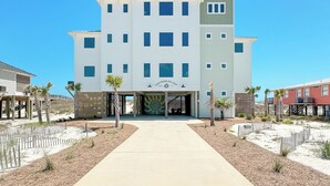 Um hotel com vários andares, entrada central, galeria coberta e paisagem arenosa com palmeiras.