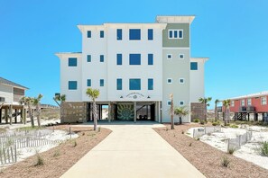 A multi-story hotel with a central entrance, a covered walkway, and a sandy landscape with palm trees.