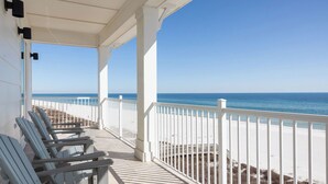 Um deck coberto com guarda-corpos brancos e cadeiras Adirondack azuis, com vista para uma praia.