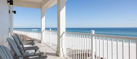 A covered deck with white railings and blue Adirondack chairs overlooking a beach.