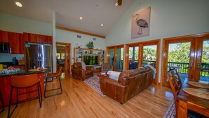 A spacious living area with a wooden floor, a large sofa, and a kitchen with a stainless steel refrigerator. - Captivating Cabana – Luxury home, Boat Slip, Golf Cart, Pool, Pickleball, & Club (North Captiva)