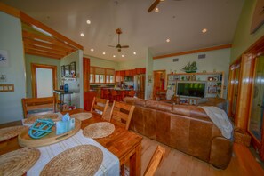 A spacious living room with a brown leather sofa, wooden dining table with placemats, and a television in the background.