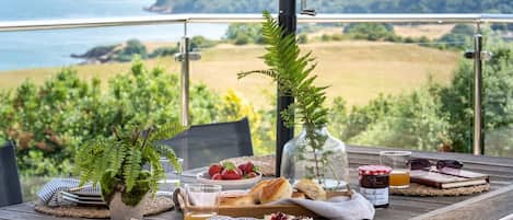 A dining table with food and drinks, a view of the sea, and a grassy landscape.
