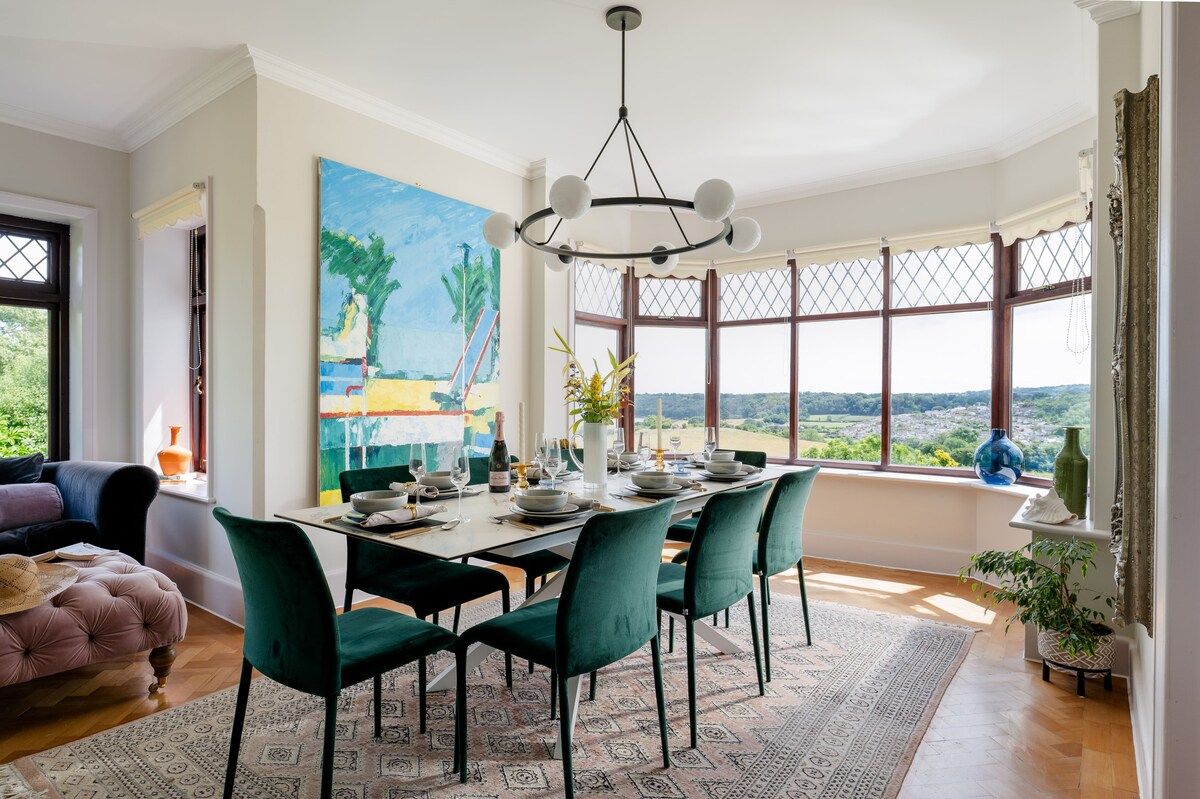 A dining area with a view, featuring a table set for six, green chairs, and a large painting on the wall.