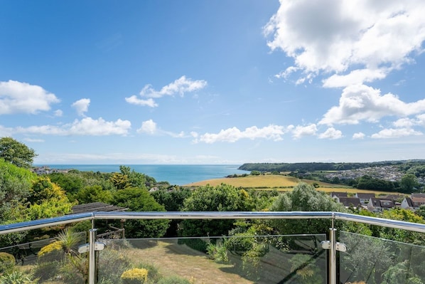 A panoramic view from a balcony with a glass railing, overlooking a coastal landscape with fields, a beach, and a distant town.