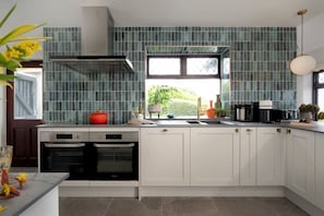 A modern kitchen with white cabinets, a stainless steel range hood, and a window with a view of greenery. - The Spinney (Paignton)