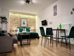 A modern room with a brick wall, a bed with green bedding, a small table with chairs, and a TV mounted on the wall. - Prosta Butelkowa (Torun)