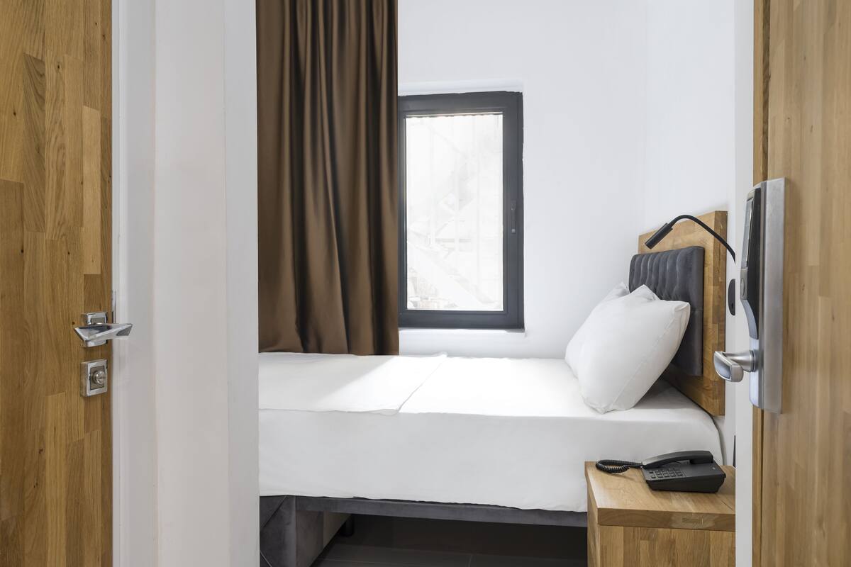 A small, single-bed room with a wooden floor, a window with curtains, a bedside table with a phone, and a wall-mounted TV.