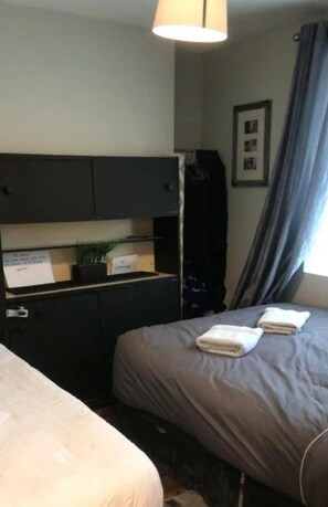 Ein Hotelzimmer mit einem Bett, einem Schreibtisch mit Computer, einem Regal mit Pflanze und einem Fenster mit Vorhängen.