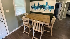 A dining area with a wooden table and white chairs, a cityscape painting on the wall, and a window with blinds.