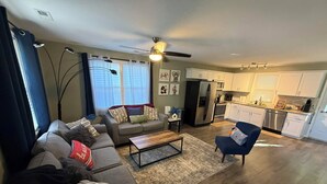 A modern living room with a sofa, armchair, and coffee table. A kitchen with appliances and cabinets is visible in the background.