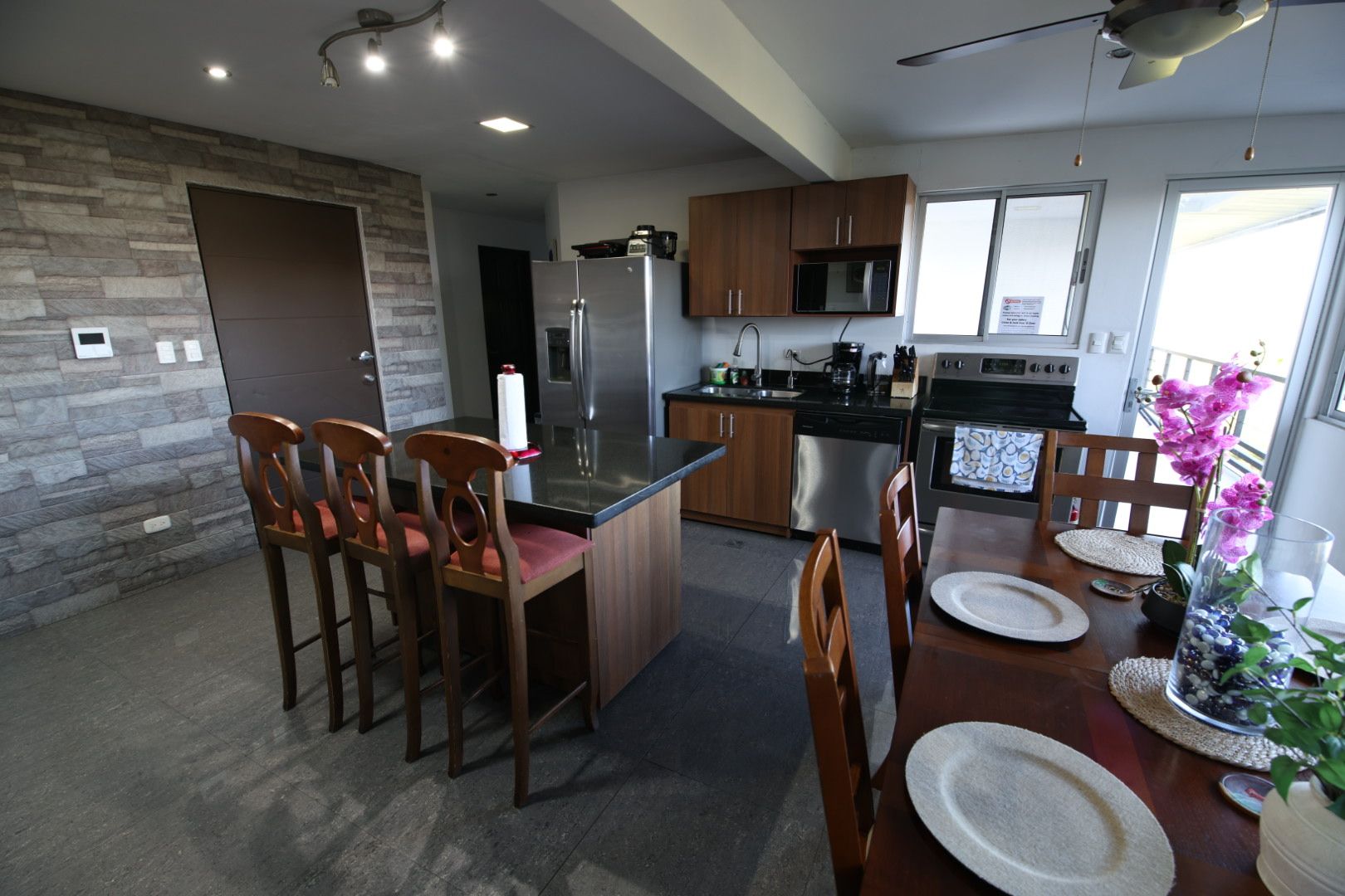 A modern kitchen with a central island, wooden chairs, a dining table with plates, and a potted plant.