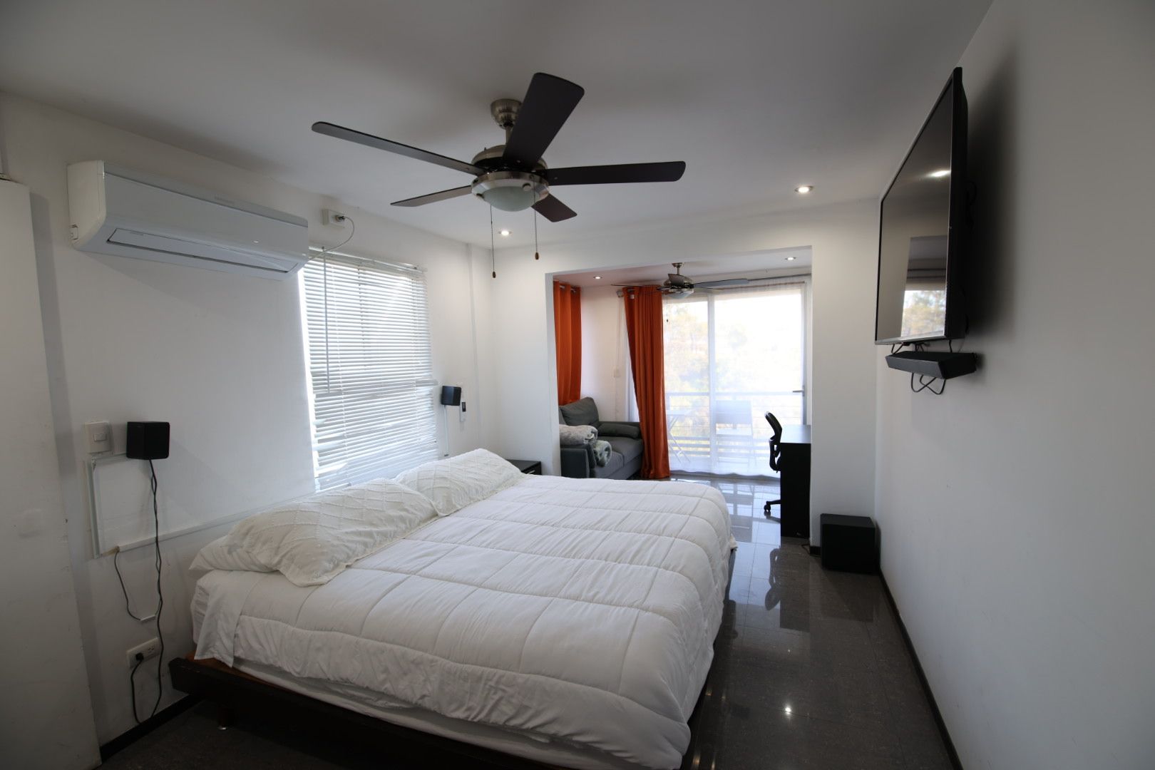 Un dormitorio moderno con cama, ventilador de techo, televisor y balcón con cortinas.