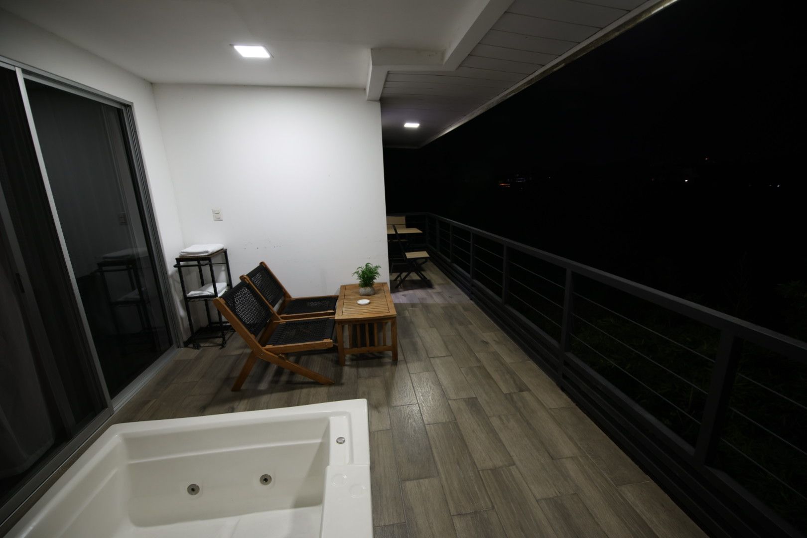 Un balcón con piso de madera, una bañera blanca, dos sillas negras, una pequeña mesa con una planta y una baranda.